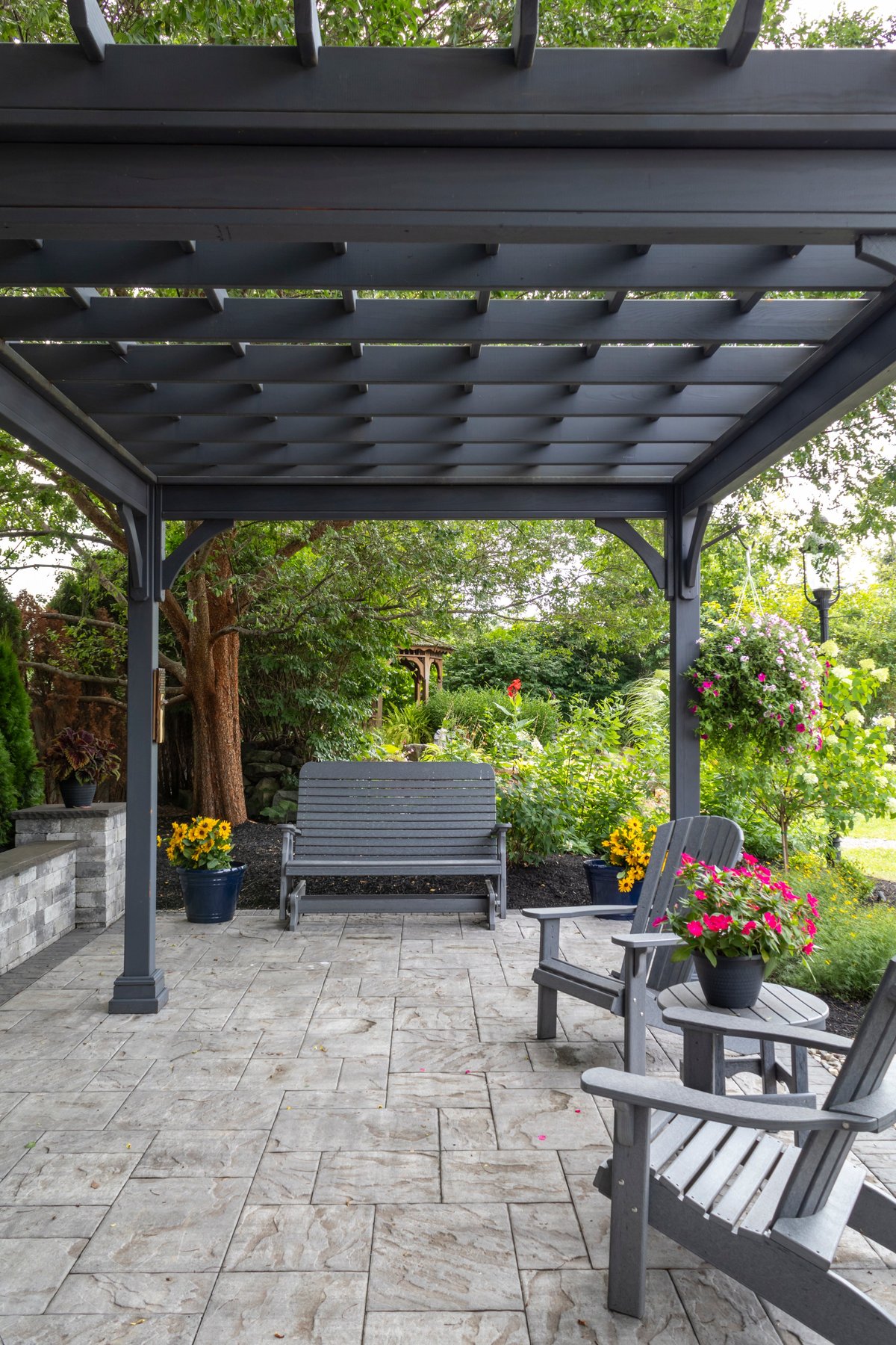 A gray wooden pergola on a paver patio with a stone fireplace and urns